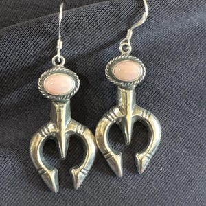 May include: A pair of silver dangle earrings featuring a polished, oval-shaped light pink stone set within a twisted rope border. Below the stone, the silver metal forms a three-pronged, crescent-like shape with etched line details.