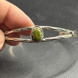 May include: A silver cuff bracelet featuring a central oval green stone set in a twisted wire bezel. The split-shank band is decorated with stamped sunburst patterns along the metal surface, creating a detailed and textured appearance.