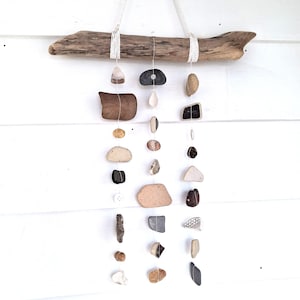 May include: A driftwood mobile featuring a collection of various shaped and colored stones suspended by thin white cords. The stones range in color from beige to dark brown and gray. The driftwood is a light brown color and is suspended by a white cord.