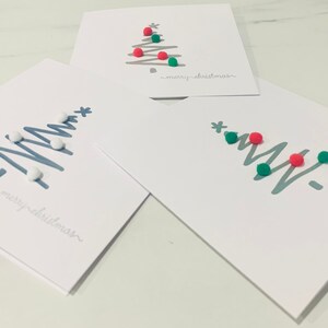 May include: Three white cards with a silver outline of a Christmas tree and pom-poms in different colours. The cards say "merry christmas" in a cursive font.