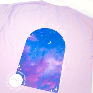 May include: A lavender long-sleeve t-shirt with a graphic of a night sky with stars and a crescent moon in an archway. The text "Zero O'Clock" is written above the archway. The text "Life whispers in twilight's breath for a little while" is written below the archway.