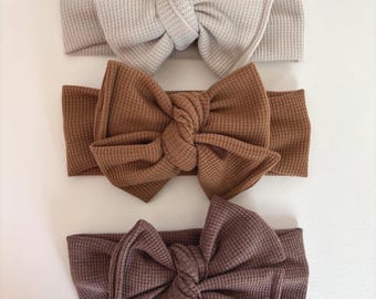 May include: Three waffle-knit headbands in neutral colors: beige, brown, and mauve. Each headband features a large, knotted bow. The headbands are arranged vertically against a white background.