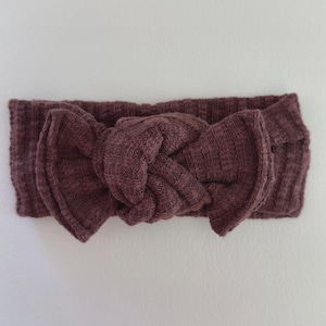 May include: A dark mauve ribbed fabric headband featuring a central knotted bow detail. The accessory is constructed from a textured, stretchy material with a consistent deep purple-brown color throughout the entire knotted design.