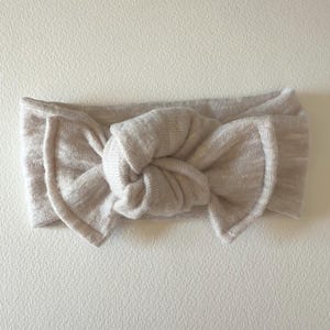 May include: A light beige headband featuring a central knotted bow design. The accessory is crafted from a soft, fine-knit fabric with a subtle texture, showcasing a continuous band that forms a decorative knot detail at the front center.
