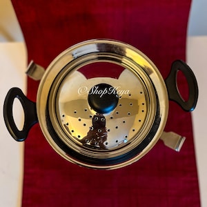 May include: A stainless steel tea infuser with a perforated lid and black handles. The infuser sits on a red runner. The text "Shop Keya" is visible on the lid.