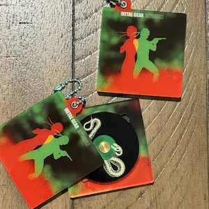 Può includere: Set custodia CD e portachiavi Metal Gear Invisible. La custodia e il portachiavi presentano un design a silhouette verde e rosso di due figure con la scritta "METAL GEAR INVISIBLE". Il CD è nero con un disegno di serpente bianco.