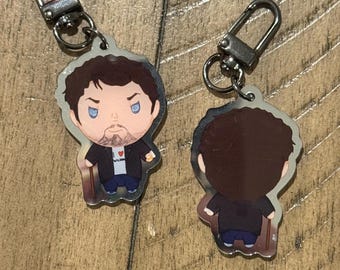 May include: Two keychains featuring a cartoon illustration of a man with a beard, wearing a black jacket, blue jeans, and a white shirt with a heart design. The keychains have silver-colored clasps.