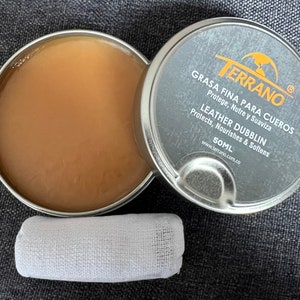 Peut inclure: Une boîte ouverte de Terrano Leather Dubblin contenant une substance cireuse brun clair, à côté d'un chiffon blanc. Le couvercle de la boîte est visible, affichant le nom de la marque et les informations sur le produit. Le produit est de 50 ml.