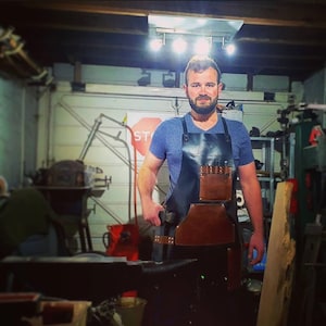 Könnte beinhalten: Ein Mann trägt ein blaues T-Shirt und eine braune und schwarze Lederschürze mit mehreren Taschen und Werkzeugschlaufen. Er steht in einer Werkstatt mit einem Amboss aus Metall im Vordergrund.