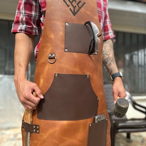 Peut inclure: Un tablier en cuir marron avec plusieurs poches, dont une grande poche sur le devant, une petite poche sur le côté et une poche pour un téléphone ou des outils. Le tablier a une boucle en métal et un logo sur le devant.