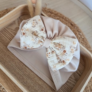 Może przedstawiać: Beżowy turban dla dziecka z dużą kokardą. Kokarda ma kwiatowy wzór w odcieniach brązu i kremu. Turban wykonany jest z miękkiej tkaniny i przeznaczony dla niemowląt.