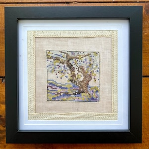 May include: A framed cross-stitch embroidery depicting a tree with purple flowers and green leaves. The embroidery is on a beige fabric and is framed in a black frame.