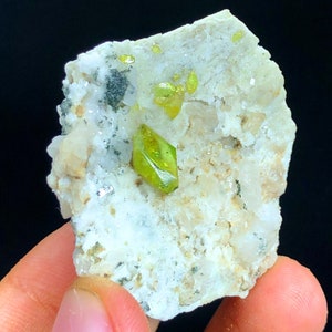 May include: A close-up of a mineral specimen featuring a light-colored matrix with embedded crystals. The crystals are a vibrant, translucent yellow-green color, with a sharp, geometric shape. The matrix has a rough, textured surface.