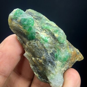 May include: A rough, gray rock with embedded green emerald crystals. The rock has a natural, unpolished look.