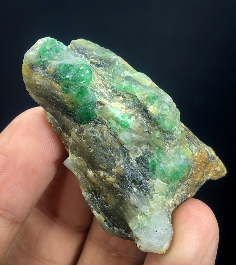 May include: A rough, gray rock with embedded green emerald crystals. The rock has a natural, unpolished look.