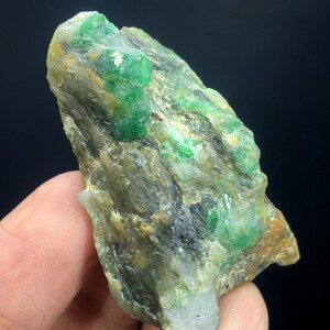 May include: A rough, gray rock with embedded green emerald crystals. The crystals are scattered throughout the rock, with some larger clusters.