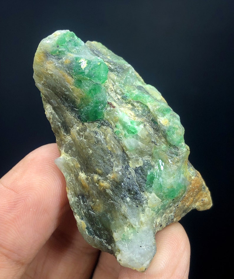 May include: A rough, gray rock with embedded green emerald crystals. The crystals are scattered throughout the rock, with some larger clusters.