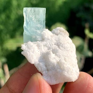 Puede incluir: Un cristal de aguamarina azul claro se encuentra sobre una base mineral blanca y texturizada. La aguamarina tiene forma rectangular con facetas visibles. El fondo es verde suave, lo que sugiere luz natural y un entorno natural.