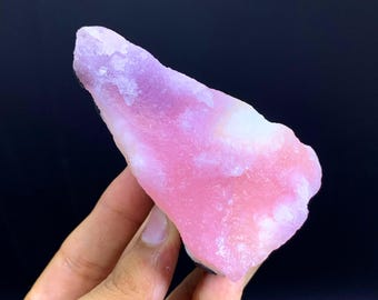 May include: A raw, uncut gemstone with a gradient of pink and lavender hues. The stone has a rough, textured surface and a natural, irregular shape. The colors blend softly, creating a visually appealing mineral specimen.