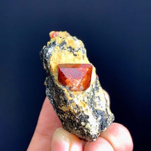 May include: A raw mineral specimen featuring a single, faceted, deep orange-red crystal embedded in a matrix of textured, dark gray and beige rock. The crystal has a distinct geometric shape with sharp, reflective edges and a smooth surface.
