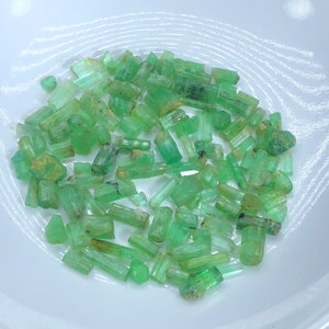 May include: A collection of raw emerald crystals in a white bowl. The crystals are a vibrant green color and have a variety of shapes and sizes.