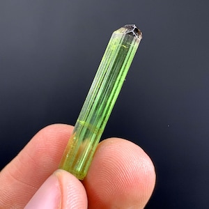 May include: A vibrant green crystal with a faceted top, held between fingers. The crystal displays natural lines and colour variations, transitioning from light to dark green, with a brown tip. The crystal is a natural mineral specimen.