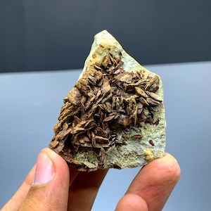 Può includere: Un esemplare di minerale naturale con una base verde chiaro e gruppi di cristalli marroni stratificati. La pietra grezza è tenuta in mano, mostrando le sue tonalità terrose e la sua consistenza unica. La superficie del minerale è ruvida e irregolare.