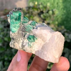 May include: A raw emerald crystal cluster with vibrant green emeralds embedded in a matrix of gray rock and white quartz. The rough, natural gemstone formation is displayed in a close-up shot.
