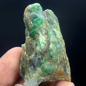 May include: A rough, green emerald gemstone embedded in a matrix of gray and brown rock. The emerald crystals are scattered throughout the rock, creating a visually interesting and unique specimen.