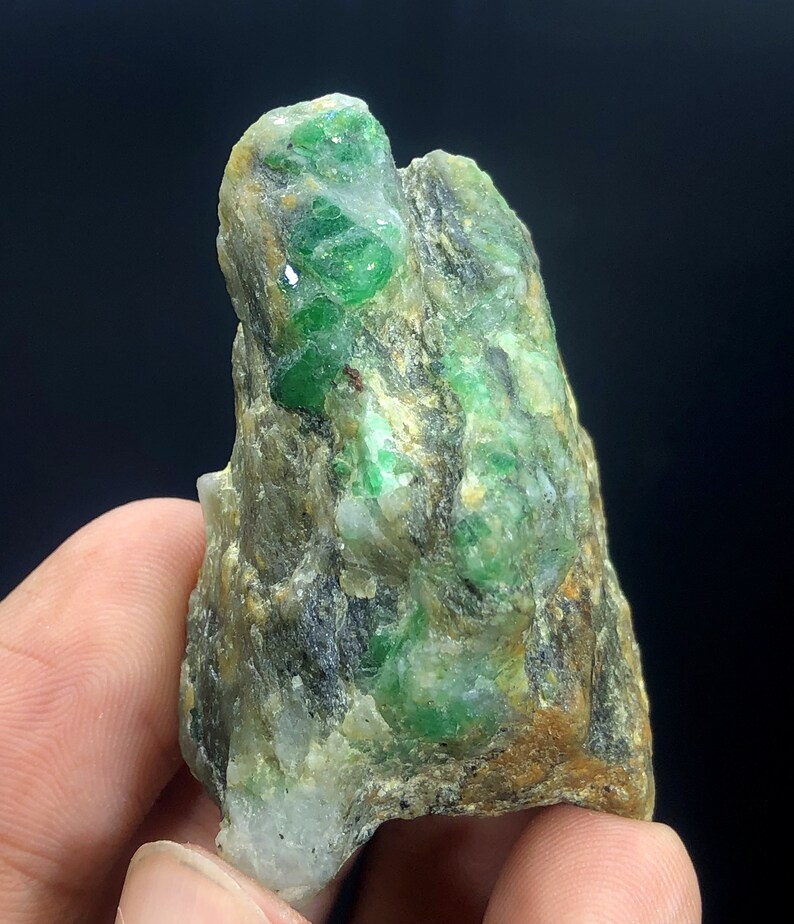 May include: A rough, green emerald gemstone embedded in a matrix of gray and brown rock. The emerald crystals are scattered throughout the rock, creating a visually interesting and unique specimen.