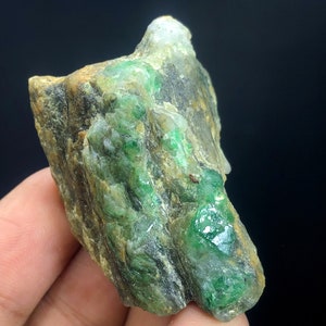 May include: A rough, green emerald gemstone embedded in a gray and brown rock matrix. The emerald has a deep green color and is surrounded by a lighter green matrix.