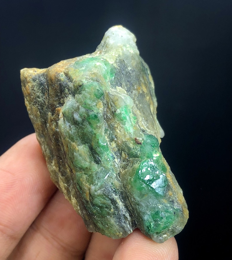 May include: A rough, green emerald gemstone embedded in a gray and brown rock matrix. The emerald has a deep green color and is surrounded by a lighter green matrix.