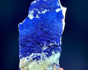 May include: A vibrant blue mineral specimen with a textured surface. The stone features a deep, saturated blue color with lighter blue and white accents. The bottom portion has a light green hue. The mineral is held in a hand against a dark blue background.