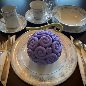 Puede incluir: Un objeto esférico morado con un diseño en espiral, que se asemeja a una fruta estilizada, se encuentra sobre un plato decorativo. El plato forma parte de una mesa formal, que incluye cubiertos, servilletas y porcelana con un estampado floral.