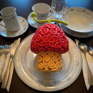 Könnte beinhalten: Eine skurrile, pilzförmige Skulptur mit roter Oberseite und gelbem Sockel, platziert auf einem Teller mit Blumenmuster. Das Gedeck umfasst Besteck, eine Teetasse, einen Sahnekännchen und eine Schüssel, alle mit passendem Blumendesign. Ein klares Glas und eine Serviette vervollständigen die elegante Anordnung.
