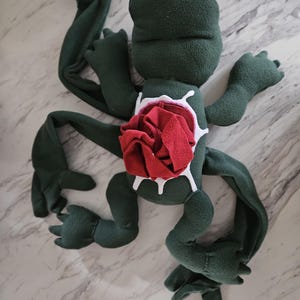 Puede incluir: Juguete de peluche verde con forma de criatura con una rosa roja en la cavidad de su pecho. La rosa está rodeada de pintura blanca que parece líquido que gotea.