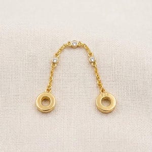 Könnte beinhalten: Ein goldfarbener Kettenverbinder mit drei kleinen, klaren, rund geschliffenen Steinen entlang der Glieder. Die Kette endet an beiden Seiten in einer flachen, runden, goldfarbenen Scheibe, wobei eine Scheibe den Stempel 925 trägt.