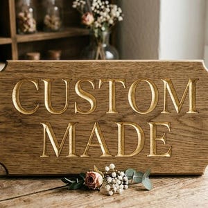 Könnte beinhalten: Ein braunes Holzschild mit den Worten "CUSTOM MADE" eingraviert. Das Schild hat einen dekorativen Rand. Eine kleine Blumengesteck befindet sich am Fuß des Schildes. Das Schild steht auf einem Holztisch. Der Hintergrund umfasst ein Fenster.