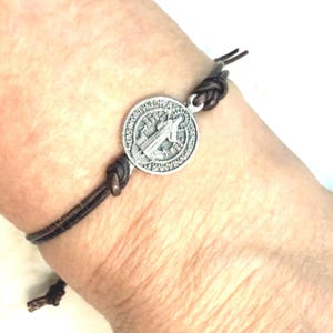 Könnte beinhalten: Ein dunkelbraunes Lederarmband mit einem silberfarbenen Medaillon. Das Medaillon hat ein geprägtes Design und ist mit Knoten am Lederband befestigt. Das Armband wird am Handgelenk getragen.
