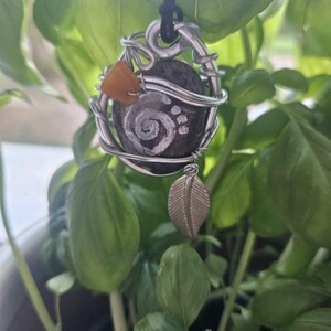 May include: A pendant featuring a dark grey stone with a white spiral and paw print design, encased in silver-toned wire wrapping. The piece includes a small amber-coloured bead and a silver-toned metal leaf charm, all suspended from a black cord.