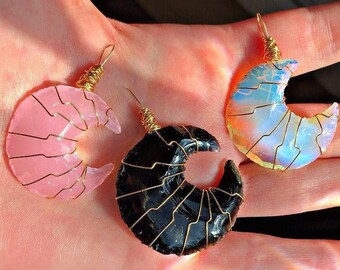 May include: Three crescent moon pendants. One is pink, one is black, and one is iridescent. Each moon is wrapped with gold-colored wire and has a loop at the top for hanging. The pendants are displayed on a light-colored background.