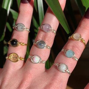 May include: A hand wearing eight different wire-wrapped rings with various gemstones. The rings are made with silver and gold wire and feature stones like amethyst, opal, and aquamarine.