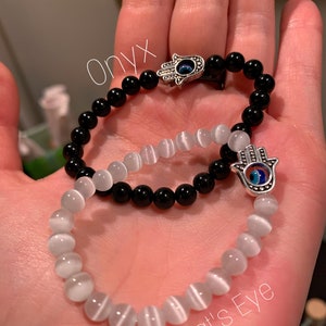 Puede incluir: Dos pulseras con dijes de mano de hamsa. Una pulsera est&aacute; hecha de cuentas de &oacute;nix negro y la otra de cuentas de ojo de gato blanco.