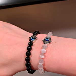 Puede incluir: Dos pulseras con dijes de mano de hamsa y cuentas de ojo turco. Una pulsera tiene cuentas negras y la otra tiene cuentas blancas.
