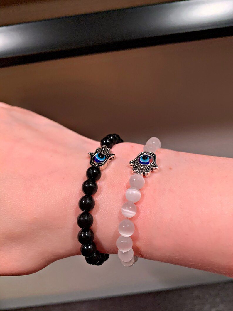Puede incluir: Dos pulseras con dijes de mano de hamsa y cuentas de ojo turco. Una pulsera tiene cuentas negras y la otra tiene cuentas blancas.