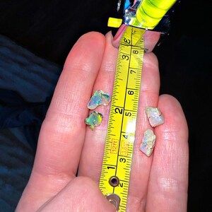 May include: A hand holding three iridescent opal gemstones, measured against a yellow tape measure. The tape measure reads 3.8 cm.