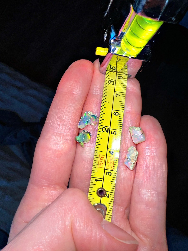 May include: A hand holding three iridescent opal gemstones, measured against a yellow tape measure. The tape measure reads 3.8 cm.