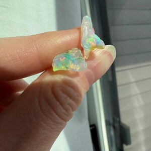 May include: Two small, irregular pieces of opal with iridescent colours, including green, blue, and yellow. The opal is translucent and has a white base.