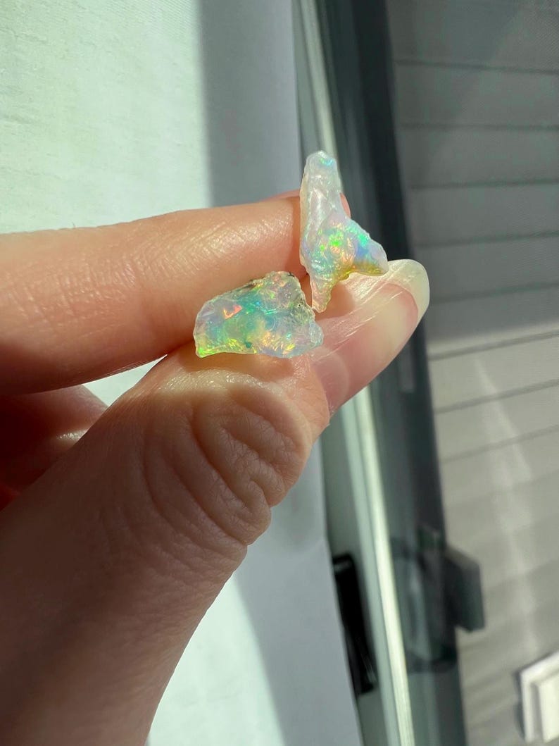 May include: Two small, irregular pieces of opal with iridescent colours, including green, blue, and yellow. The opal is translucent and has a white base.