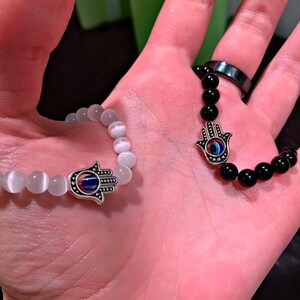 Puede incluir: Dos pulseras con dijes de mano de hamsa. Una pulsera tiene cuentas blancas y la otra tiene cuentas negras. Ambos dijes tienen un ojo turco azul en el centro.